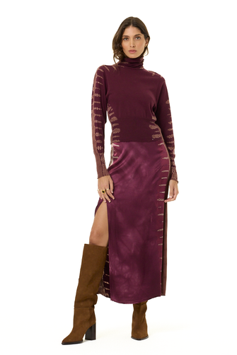A model wearing a long-sleeve turtleneck and matching skirt in a deep plum purple, featuring tie-dye details on the sleeves and sides with a high slit, paired with brown knee-high boots against a plain white background.