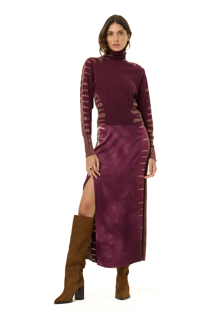 A model wearing a long-sleeve turtleneck and matching skirt in a deep plum purple, featuring tie-dye details on the sleeves and sides with a high slit, paired with brown knee-high boots against a plain white background.