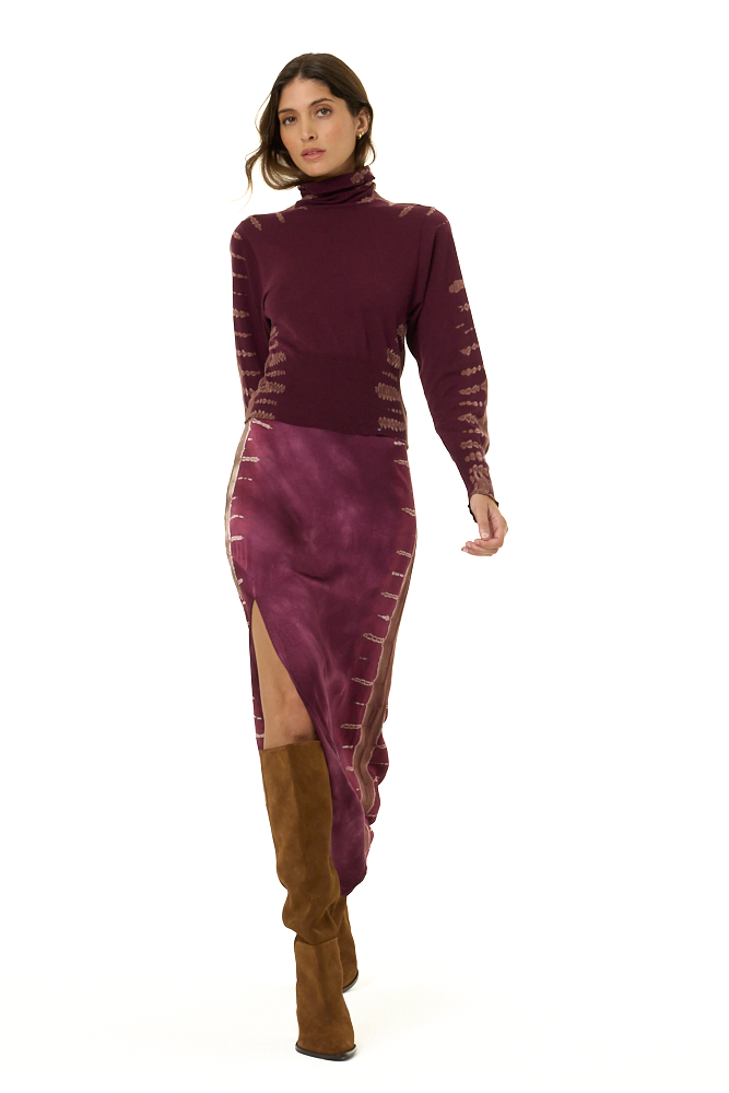 A model wearing a long-sleeve turtleneck and matching skirt in a deep plum purple, featuring tie-dye details on the sleeves and sides with a high slit, paired with brown knee-high boots against a plain white background.