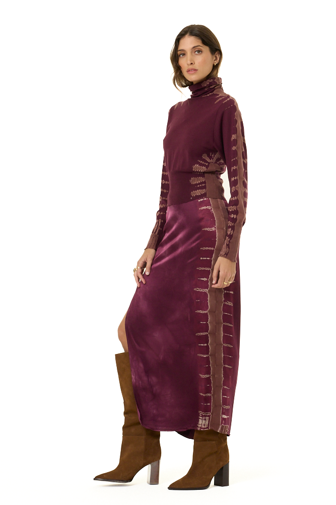 A model wearing a long-sleeve turtleneck and matching skirt in a deep plum purple, featuring tie-dye details on the sleeves and sides with a high slit, paired with brown knee-high boots against a plain white background.