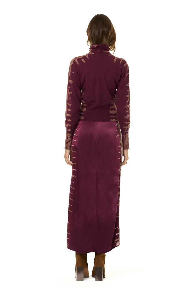 The backside of a model wearing a long-sleeve turtleneck and matching skirt in a deep plum purple, featuring tie-dye details on the sleeves and sides with a high slit, paired with brown knee-high boots against a plain white background.