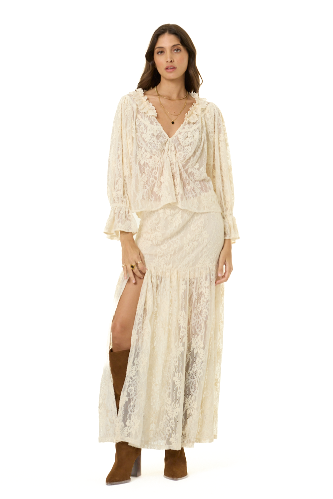 A woman modeling a cream lace two-piece outfit consisting of a long-sleeve ruffled blouse and a matching high-slit maxi skirt, paired with brown suede boots, against a solid white background.