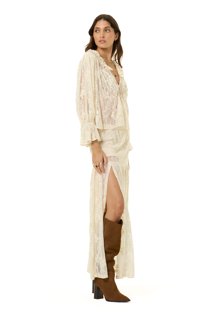 A woman modeling a cream lace top and matching maxi skirt with a high leg slit, paired with brown suede boots, against a solid white background.