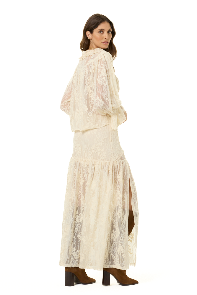 A woman modeling a cream lace blouse and matching maxi skirt with long sleeves, and a tiered skirt with side slits, paired with brown ankle boots, against a plain white background.