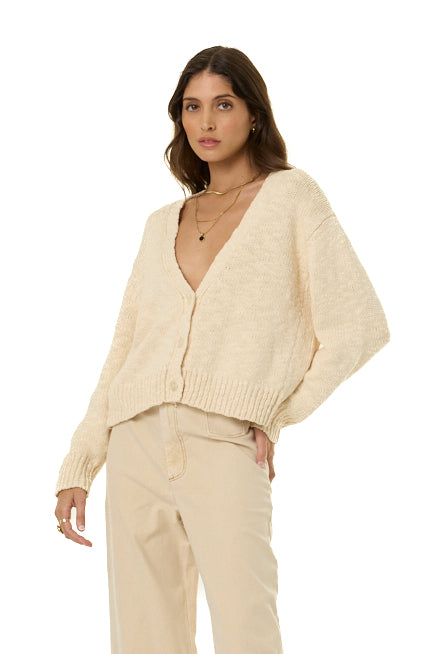 A woman modeling a cream-colored, textured knit cardigan sweater with a deep V-neck and button-front closure, paired with matching cream trousers, against a solid white background.