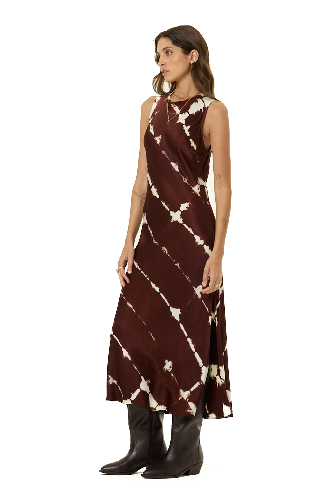 A woman modeling a sleeveless, high-neck midi dress with a brown and cream tie-dye pattern, paired with dark brown leather boots, set against a plain white background.