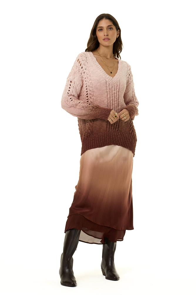 A woman modeling a pink and brown gradient knit sweater dress with a chunky knit V-neck top and flowing chiffon and satin layered skirt, paired with black boots, on a white background.