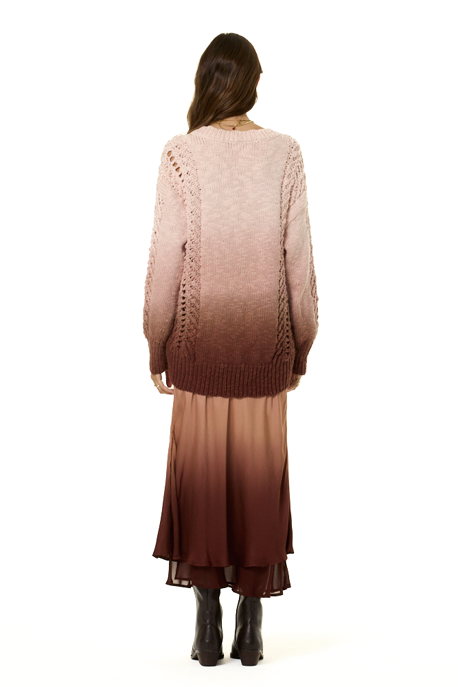 A woman viewed from behind wearing a long-sleeve knit sweater with an ombré gradient from light blush pink at the top to deep brown at the bottom, paired with a matching ombré midi skirt and black ankle boots, against a plain white background.