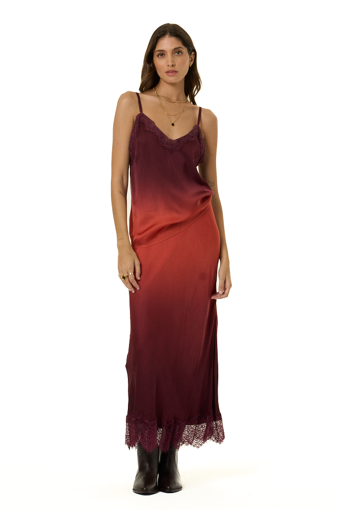 A woman modeling a long satin cami and skirt set with a deep burgundy-to-terracotta red ombré gradient and lace trim at the hem, paired with black boots against a plain white background.