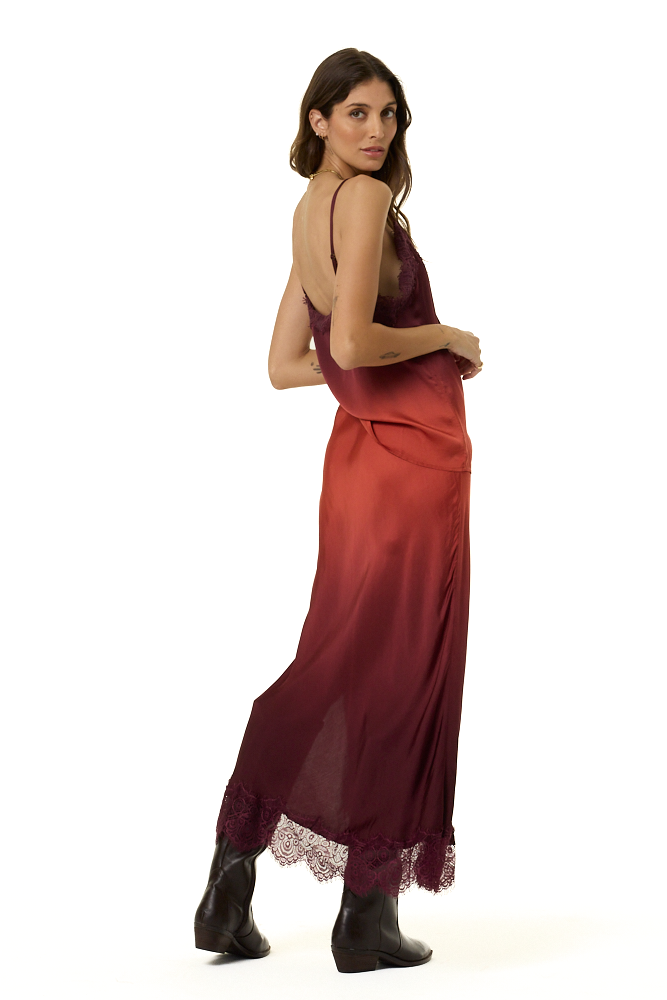 A woman modeling a long, sleeveless satin cami and skirt set with a gradient color story, fading from deep burgundy at the top to a muted rust red at the hem, styled with black ankle boots against a plain white background.