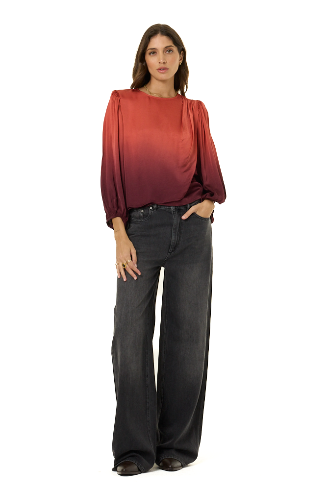 A woman modeling a long-sleeve satin top with a red-to-orange ombré gradient, paired with dark gray wide-leg jeans, against a solid white background.