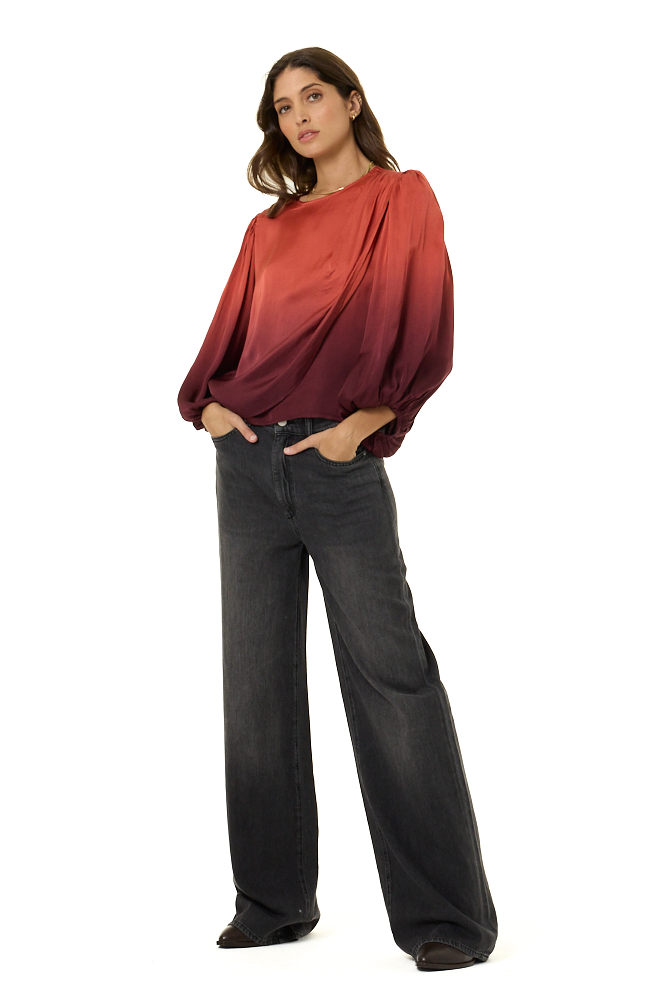 A woman modeling a long-sleeve draped top with a red-to-orange gradient, paired with dark gray wide-leg jeans, against a solid white background.