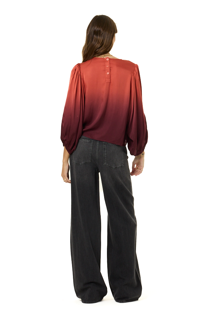 A woman viewed from the back wearing a long-sleeve satin-like top with a red-to-burgundy ombré gradient and wide-leg dark gray pants, against a plain white background.