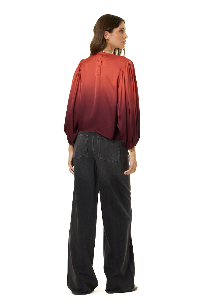 A woman shown from the back wearing a long-sleeve satin-like top with a red-to-dark-burgundy ombre gradient and wide-leg dark gray pants, photographed against a plain white studio background.