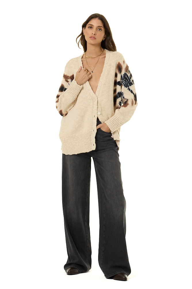 A woman modeling a cream-colored knit cardigan with brown and black leopard print sleeves and a blue patterned yoke, paired with dark wide-leg jeans against a plain white background.