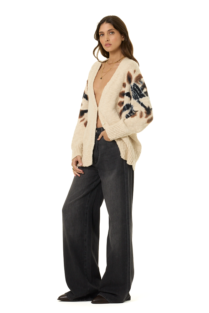 A woman modeling a cream knit cardigan with brown and black abstract patterns on the sleeves, paired with dark wide-leg jeans, against a plain white background.
