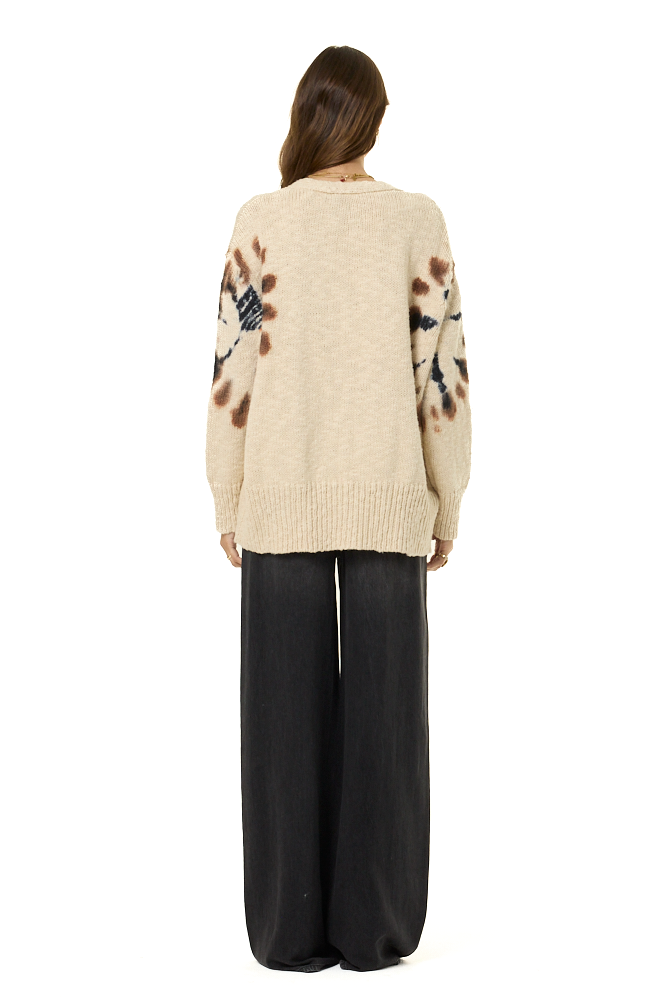 A woman viewed from the back wearing a cream-colored knit sweater with brown and black abstract patterns on the sleeves, paired with black wide-leg pants, against a plain white background.