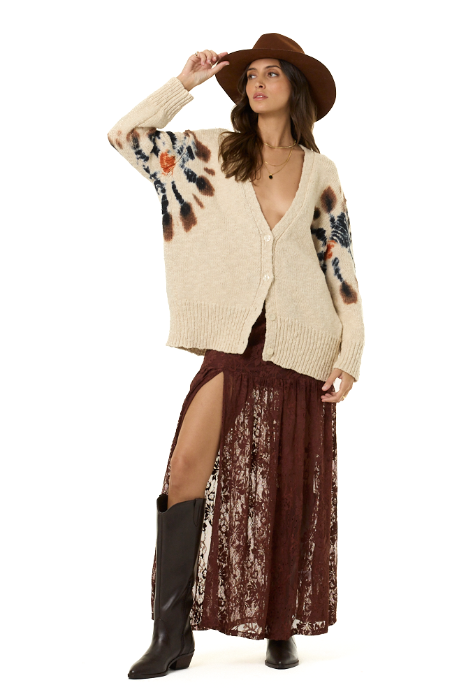 A woman modeling a cream knit cardigan with brown and orange spiral-like patterns on the sleeves, paired with a brown wide-brim hat, a dark brown lace maxi skirt, and brown boots against a white studio background.
