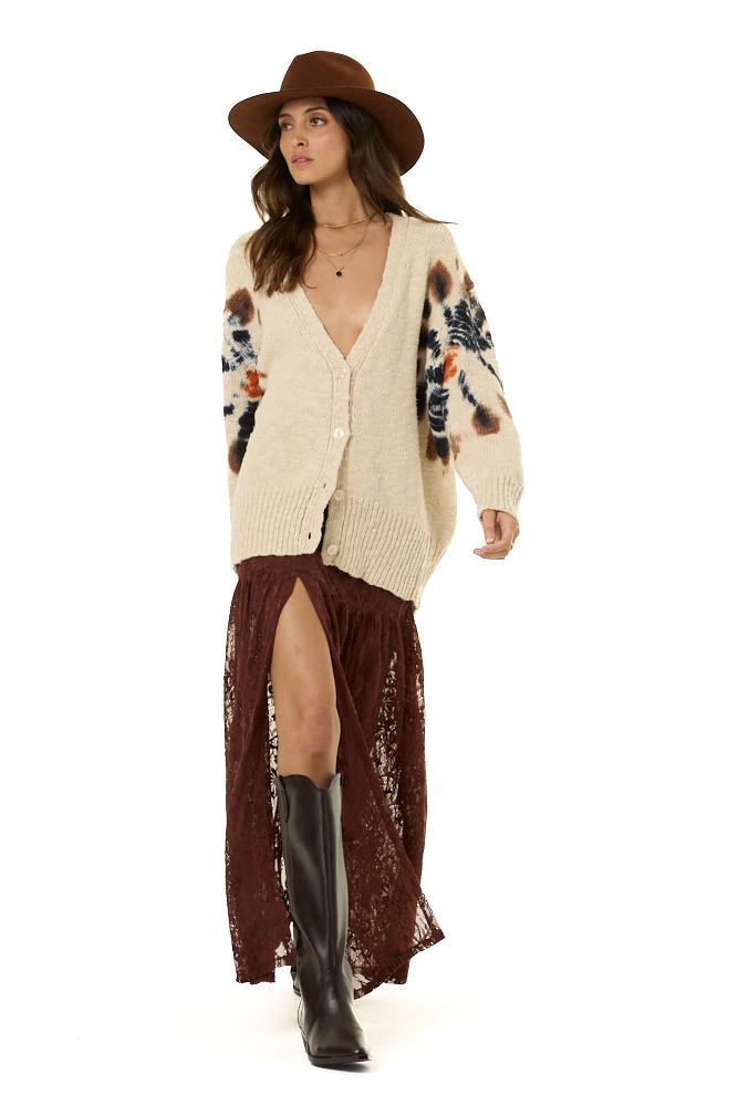 A woman modeling a cream knit cardigan with brown and orange spiral-like patterns on the sleeves, paired with a brown wide-brim hat, a dark brown lace maxi skirt, and brown boots against a white studio background.