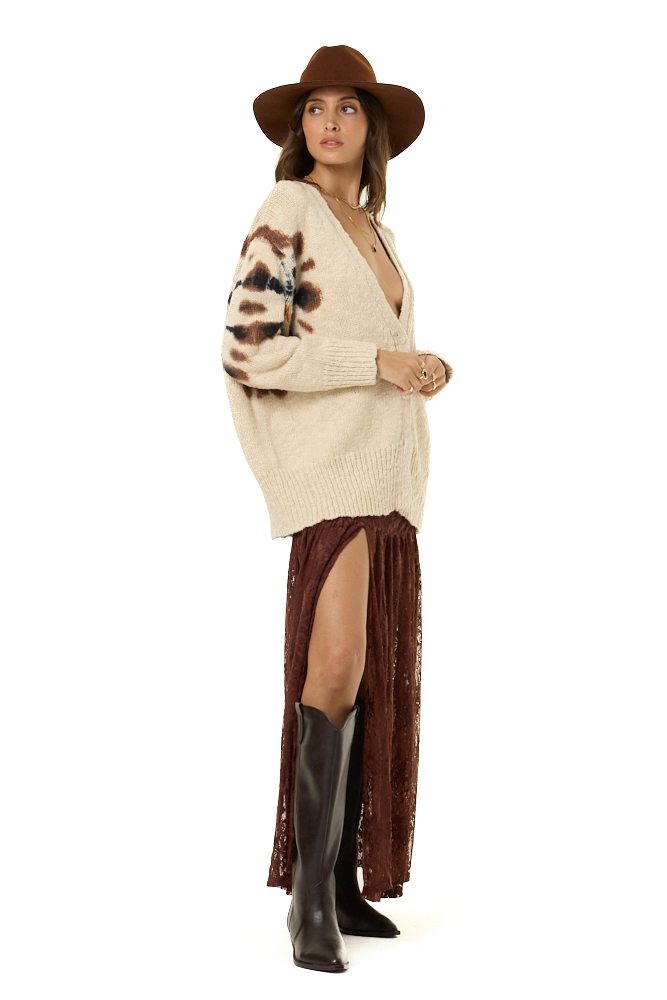 A woman modeling a cream knit cardigan with brown abstract patterns on the sleeves, paired with a brown wide-brim hat, a brown lace skirt, and brown boots against a white background.