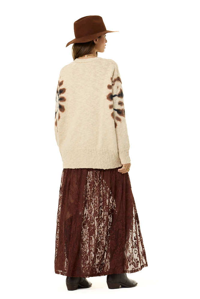 A model shown from the back wearing a cream-colored knit sweater with brown sunburst patterns on the shoulders, a dark brown lace maxi skirt, a brown wide-brim hat, and brown boots, set against a plain white background.
