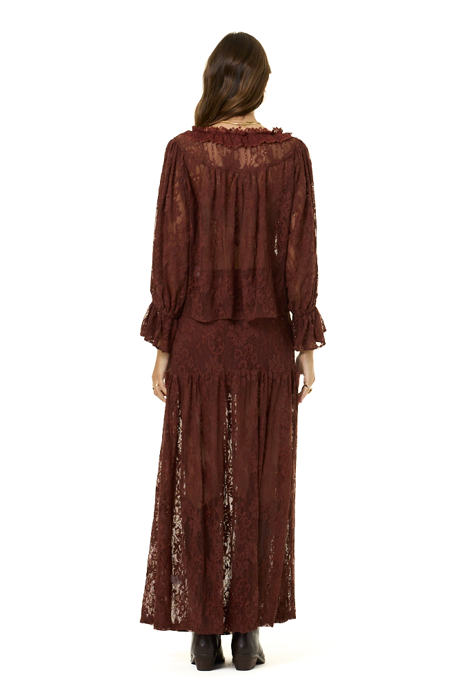 A woman viewed from the back wearing a long-sleeved, dark brown lace blouse and matching maxi skirt with a ruffled neckline and a matching skirt, paired with black boots, against a solid white background.