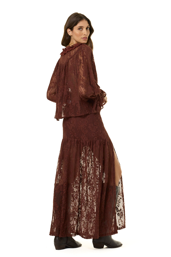 A woman modeling a dark brown lace two-piece outfit consisting of a long-sleeve top and a matching maxi skirt, shown from a three-quarters back view against a white background.