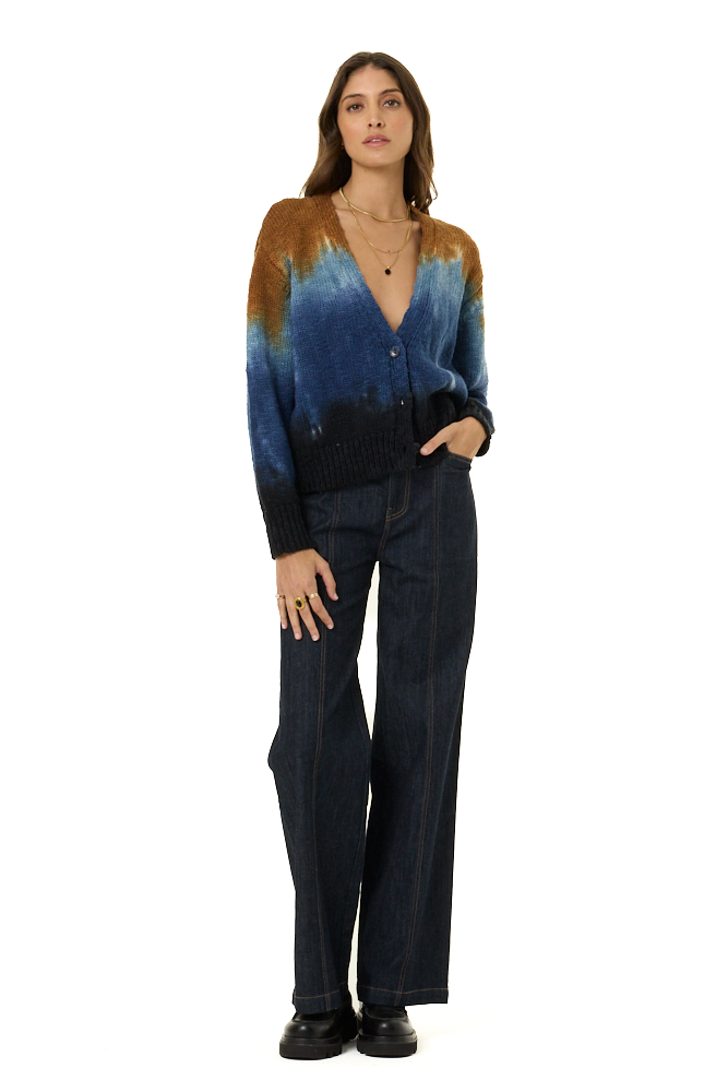 A woman stands against a white studio background wearing a long-sleeve knit cardigan with a blue, teal, and brown gradient pattern, paired with dark blue wide-leg jeans and black shoes.