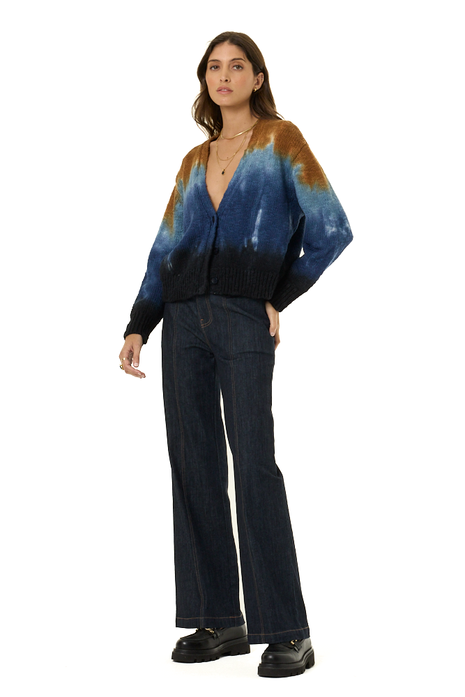 A woman modeling a tie-dye cardigan with blue, brown, and black patterns, paired with dark blue wide-leg jeans and black loafers, against a solid white background.