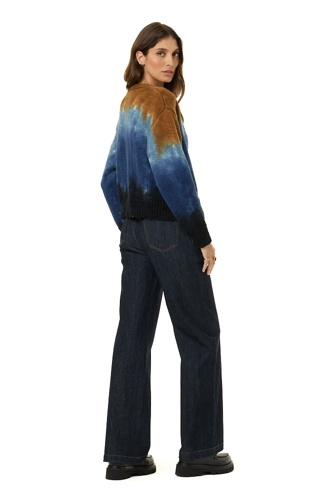 A woman modeling a blue and brown tie-dye knit sweater with dark wide-leg jeans and black shoes, shown from a three-quarters back view against a plain white background.