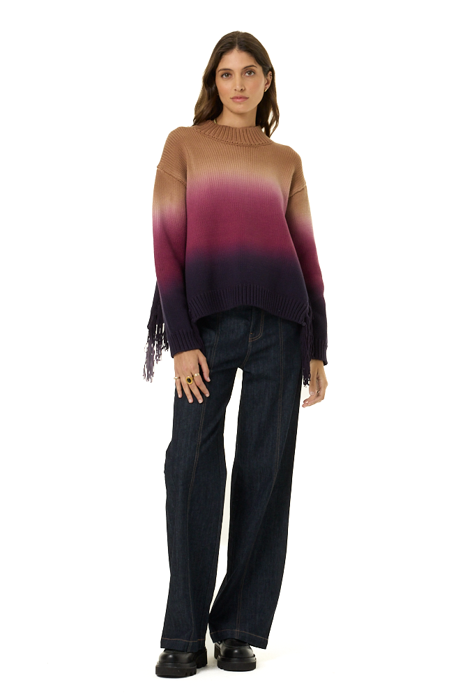 A woman modeling a loose-fit knit sweater with a vertical ombré color-block pattern transitioning from tan/beige at the top, through magenta and purple, to a very dark plum at the bottom. The sweater features fringe detailing on one sleeve.