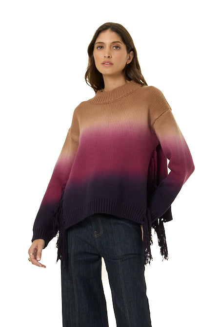 A woman modeling a long-sleeve knit sweater with a brown, magenta, and dark purple ombre gradient, featuring fringe details along the hem and cuffs, paired with dark blue jeans against a white background.