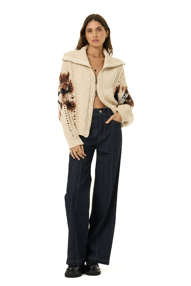 A model stands against a white studio background wearing a cream-colored knit zip-up jacket with brown and black patterned sleeves, paired with dark wide-leg jeans and black shoes.