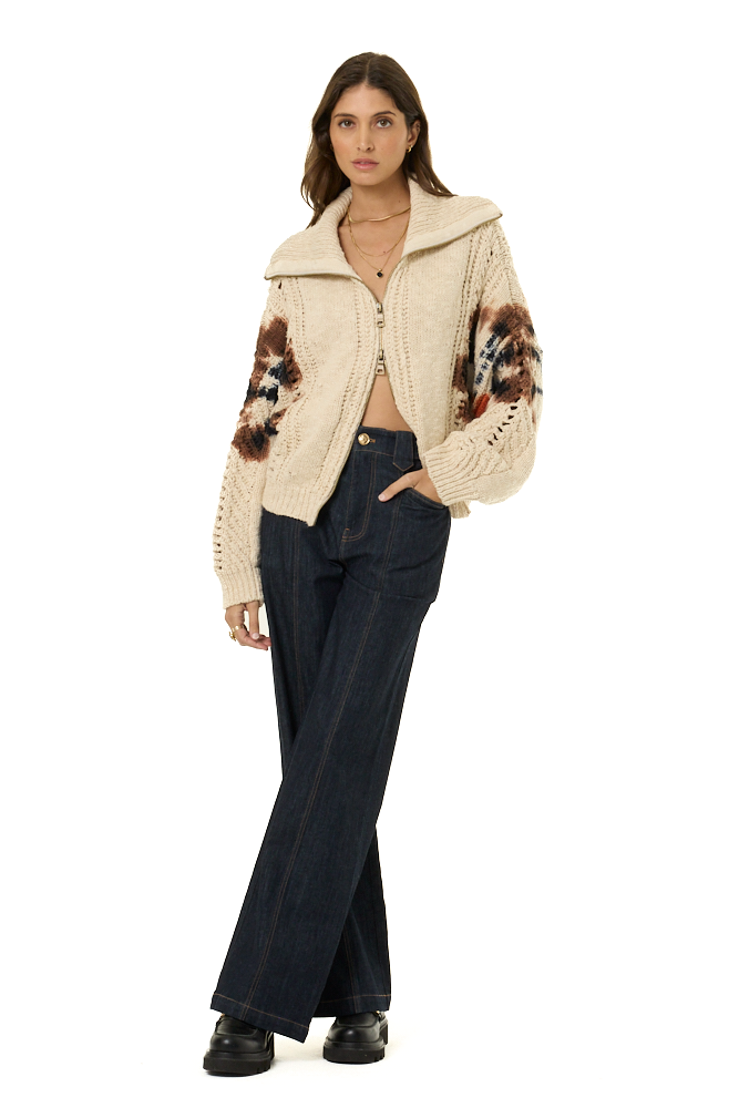 A woman modeling a cream cable-knit cropped zip-up hoodie with brown and black patterned on the sleeves, paired with dark blue wide-leg jeans and black shoes, against a solid white background.