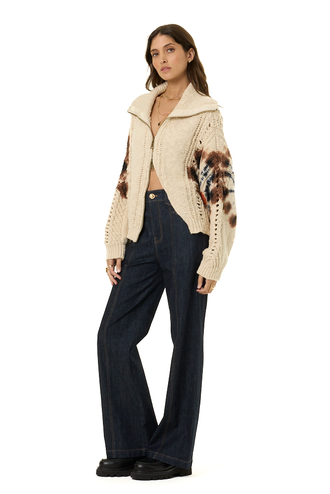 A woman modeling a cream cable-knit zip-up cardigan with brown, black, and white patterned sleeves, paired with dark blue wide-leg jeans and black shoes, against a solid white background.