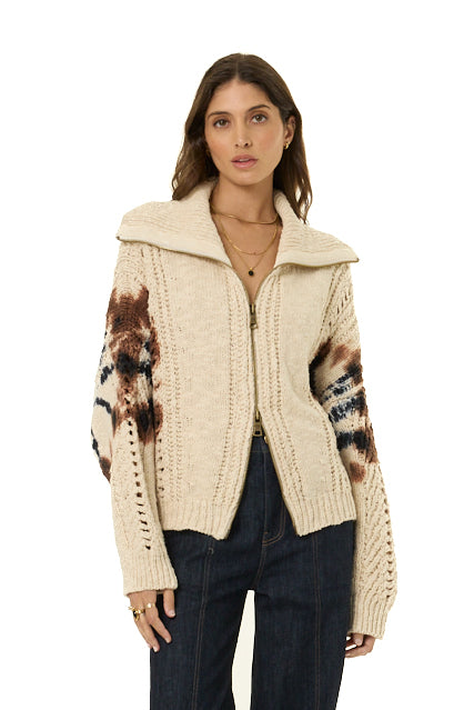 A woman modeling a cream-colored cable-knit zip-up cardigan with brown and black patterned sleeves, paired with dark blue jeans, against a plain white background.