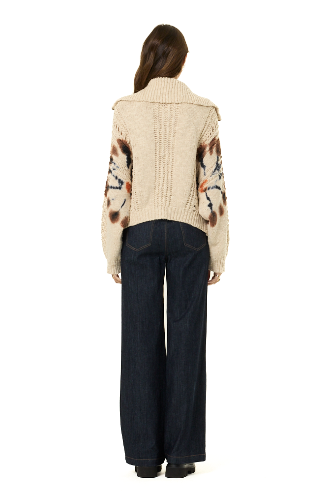 A woman viewed from the back wearing a cream cable-knit cropped sweater with tie-dye pattern on sleeves, paired with dark wide-leg jeans and black shoes, against a plain white background.