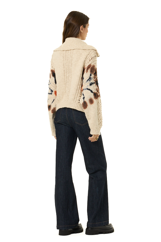A woman shown from the back wearing a cream cable-knit cropped sweater with brown and black patterns on the sleeves, paired with dark blue jeans and black shoes, against a plain white background.