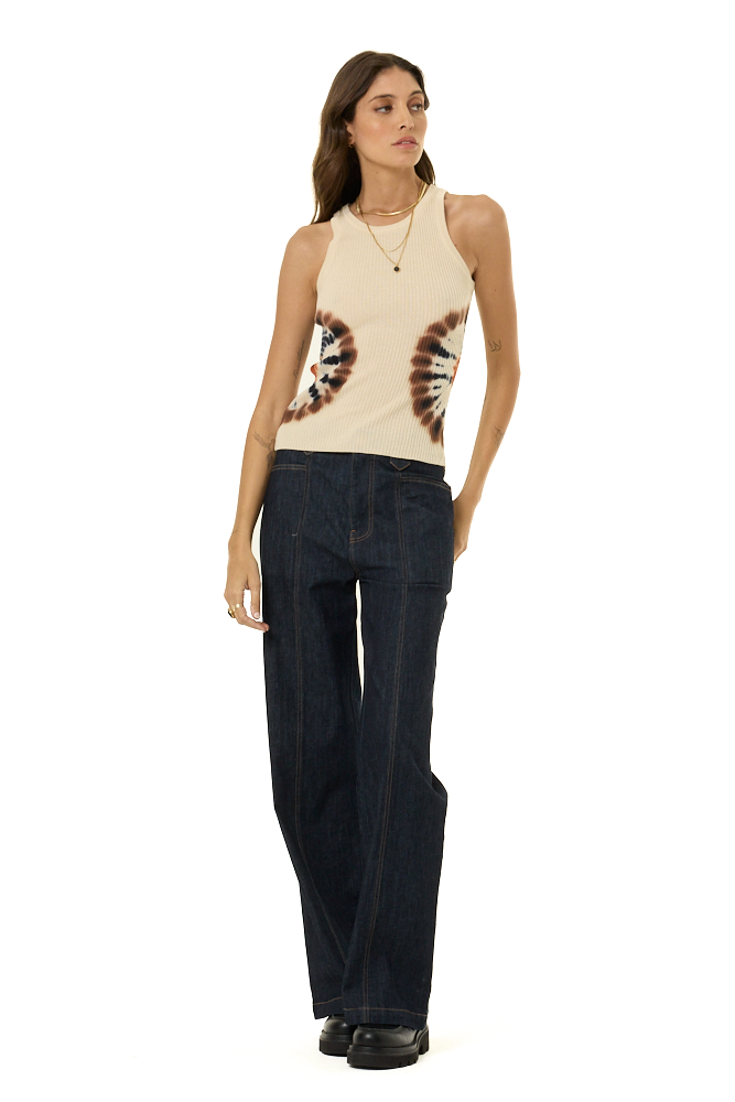 A woman modeling a sleeveless cream sweater tank top with brown and black tie-dye circular patterns on the sides, paired with dark wide-leg jeans and black shoes, against a plain white background.