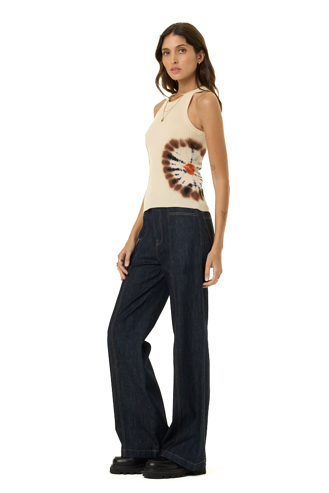 A woman modeling a sleeveless cream sweater tank top with a brown and white tie-dye circular graphic on the front, paired with dark wide-leg jeans and black shoes, against a plain white background.