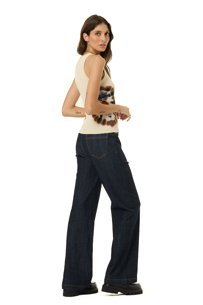 A woman modeling dark wash wide-leg jeans and a cream sleeveless sweater tank top with a brown graphic print, shown from a three-quarters back angle against a plain white background.