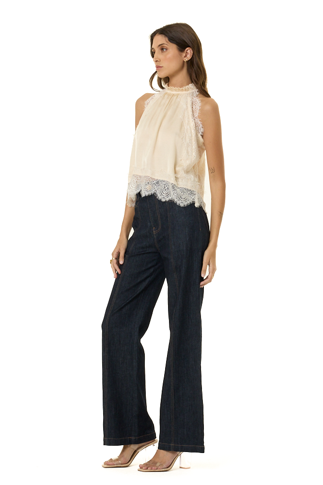 A woman modeling a sleeveless cream top with lace trim and dark wide-leg jeans against a plain white background.