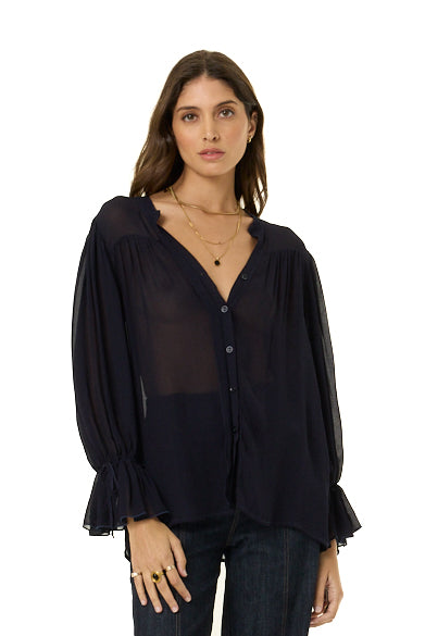 A woman modeling a sheer navy long-sleeve blouse with a V-neckline, button front, and ruffled cuffs, paired with dark jeans against a plain white background.