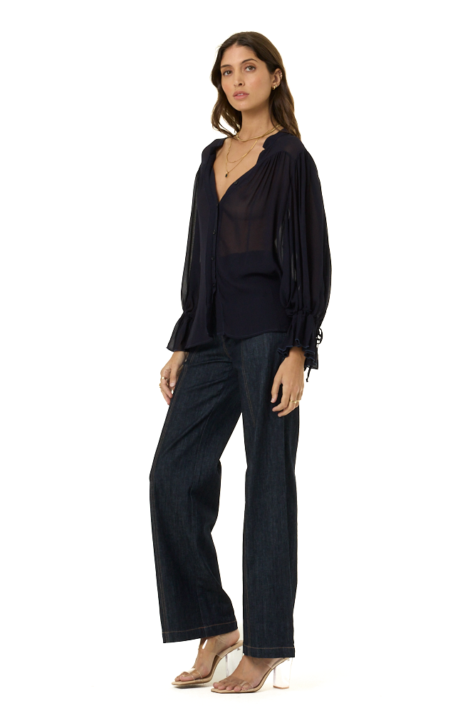A woman modeling a dark navy blue outfit consisting of a draped long-sleeve top, a sheer navy V-neck top, dark wide-leg pants, and clear high-heeled sandals, set against a plain white background.