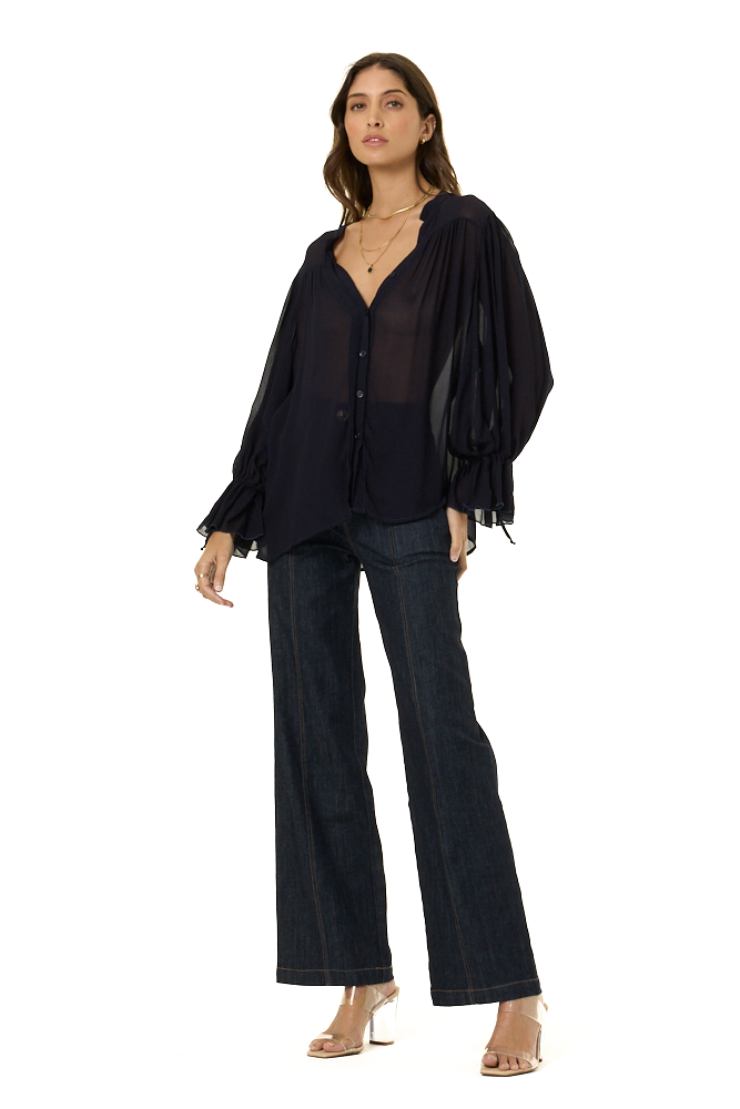 A woman modeling a navy sheer long-sleeve blouse with dramatic puffed sleeves and dark wide-leg jeans against a plain white background.