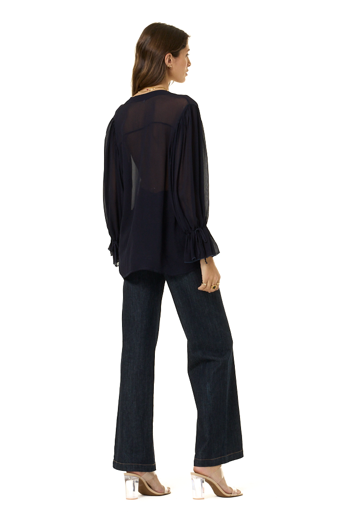 A woman shown from the back wearing a dark navy sheer long-sleeve blouse with gathered ruffle sleeves and dark wide-leg pants, photographed against a plain white studio background.