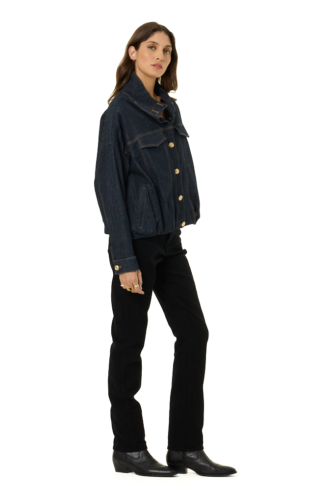 A woman modeling a dark indigo denim jacket with gold-tone buttons, paired with black pants and black leather boots, shown in profile against a solid white background.