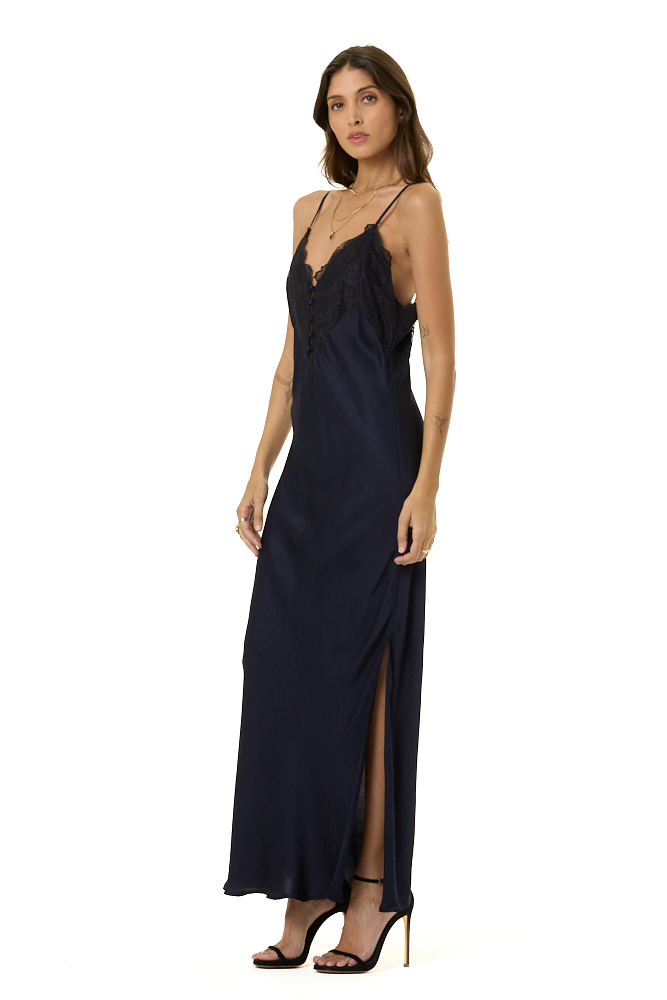 A woman modeling a long navy blue satin slip dress with thin straps, a plunging lace neckline, and a high side slit, paired with black strappy heels against a plain white background.