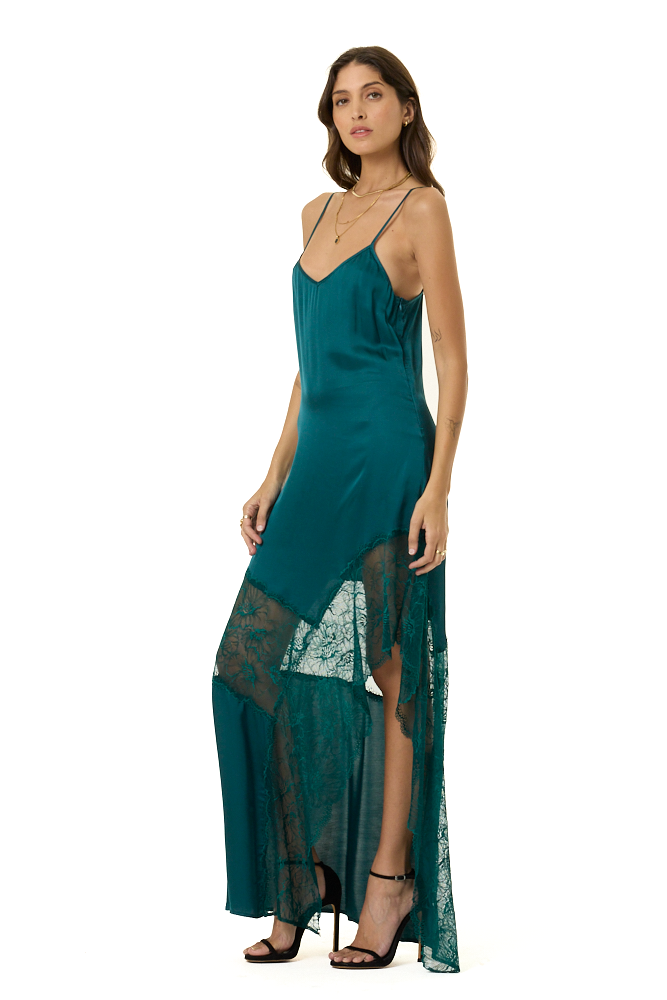 A woman modeling a long teal slip dress with sheer lace panels and a high slit, paired with black strappy heels, against a plain white background.
