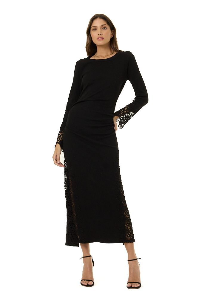 A woman modeling a black long-sleeve midi dress with sheer lace trim on the cuffs and side seams, paired with black strappy heels, against a plain white background.